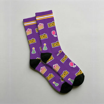 Calcetines personalizados morados con el nombre LUCÍA repetido en amarillo, entre tartas, regalos, globos y gorros de fiesta